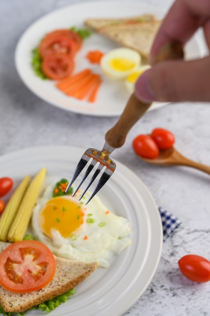 Huevos fritos, pan, zanahorias y tomates en un plato blanco para el desayuno, enfoque selectivo de mano con un tenedor.