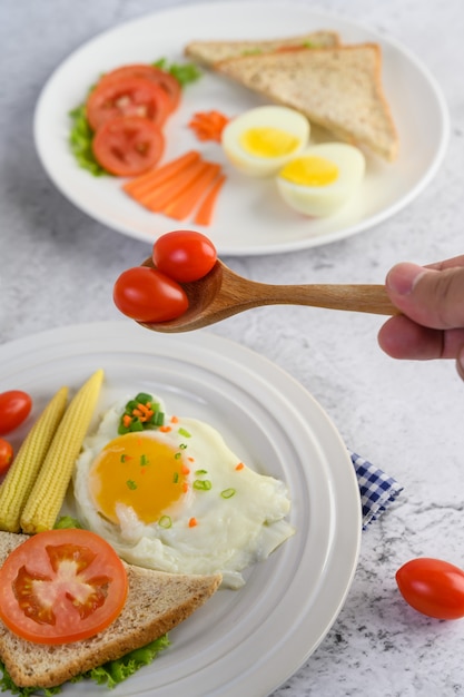 Huevos fritos, pan, zanahorias y tomates en un plato blanco para el desayuno, enfoque selectivo de mano con tomates en una cuchara de madera.
