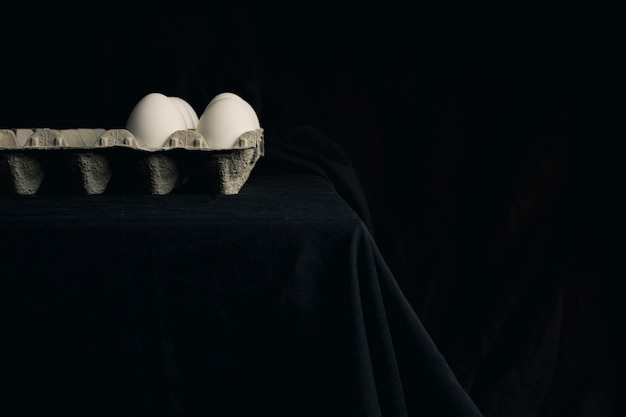 Huevos de gallina en un recipiente en el borde de la mesa entre la negrura