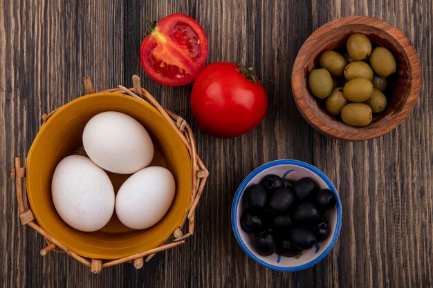 Huevos de gallina de vista superior en canasta con aceitunas negras y verdes en tazones y tomates sobre fondo de madera