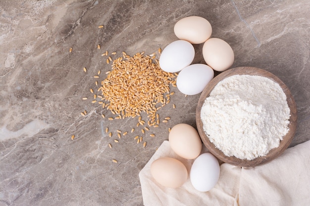 Huevos, granos de trigo y una taza de trigo triturado sobre mármol.