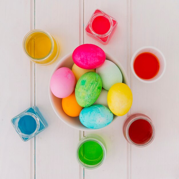 Huevos de Pascua brillantes en un tazón entre latas de líquido de tinte en el escritorio