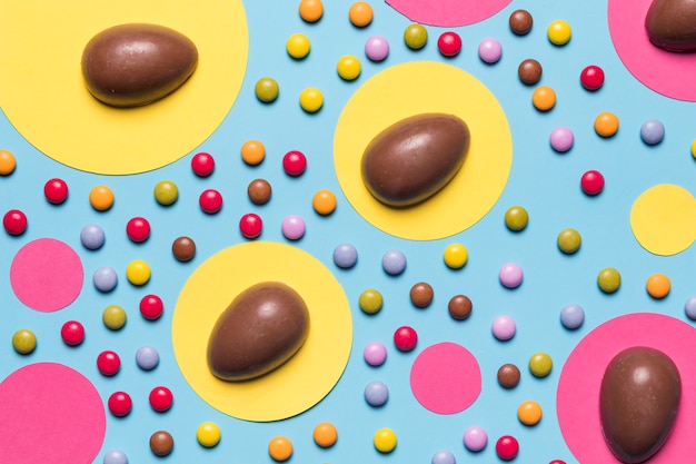 Huevos de Pascua del chocolate en el marco de papel circular rosado y amarillo adornado con los caramelos de la gema en fondo azul