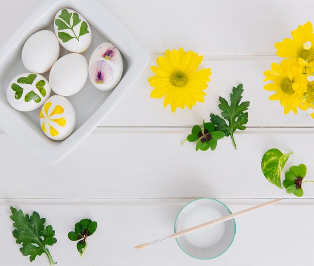 Huevos de Pascua en un recipiente cerca de flores, hojas y taza con líquido tinte