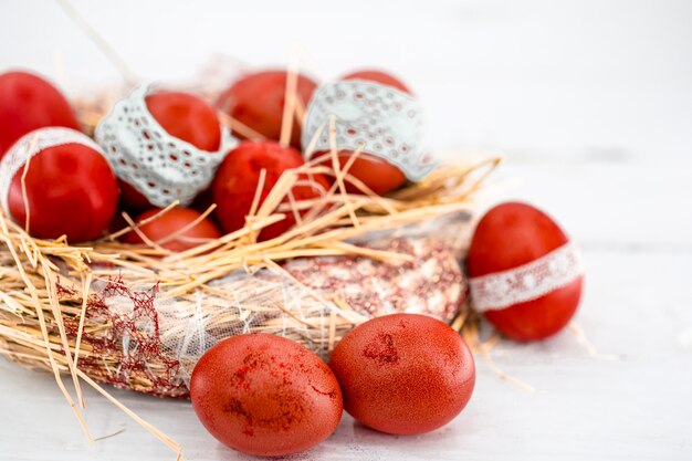 Huevos de Pascua rojos en un nido de heno, atado una cinta de encaje, de cerca, acostado sobre una madera blanca