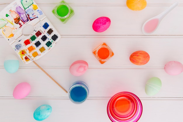 Huevos de Pascua entre tazas con líquido de tinte cerca de cucharas y pincel con un conjunto de colores