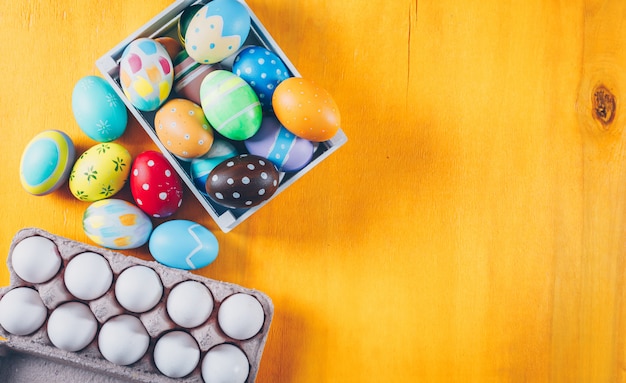 Huevos de Pascua de la vista superior en caja de madera y cartón de huevos en fondo de madera amarillo.