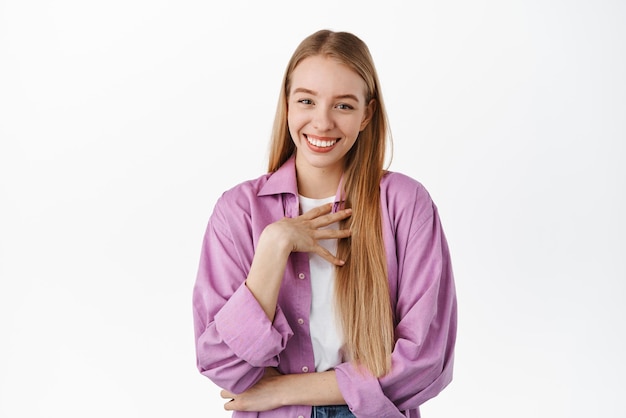 Humilde y linda chica rubia que parece tocada y halagada sonriendo complacida agradeciendo algo de pie con una elegante camisa morada contra fondo blanco