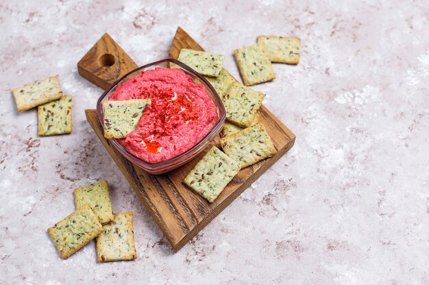 Hummus de remolacha en tabla de cortar con galletas saladas en superficie clara