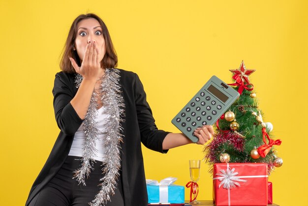 Humor navideño con una bella dama nerviosa sorprendida de pie en la oficina y sosteniendo la calculadora en la oficina en amarillo