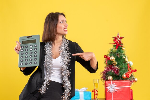 Humor navideño con hermosa dama emocional de pie en la oficina y señalando la calculadora en la oficina en amarillo
