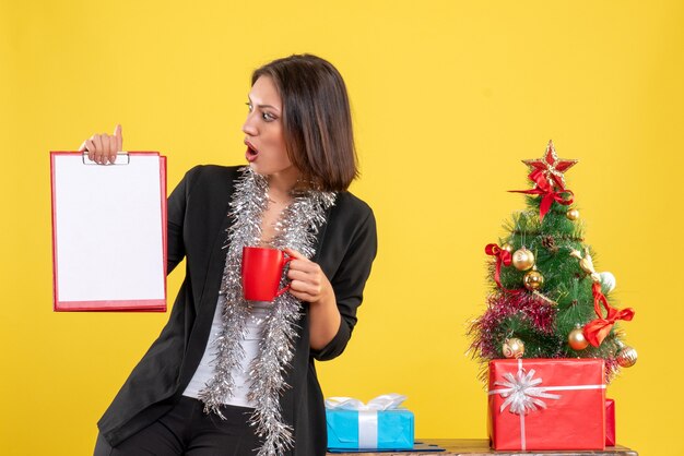 Humor navideño con hermosa dama sorprendida de pie en la oficina y sosteniendo la taza de documentos en la oficina en amarillo