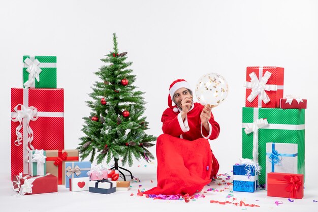 Humor navideño con joven santa claus divertido sentado cerca del árbol de navidad y regalos en diferentes colores sobre fondo blanco.
