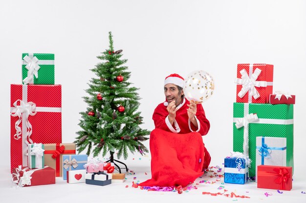 Humor navideño con joven santa claus sosteniendo globo sentado cerca del árbol de navidad y regalos en diferentes colores sobre fondo blanco.