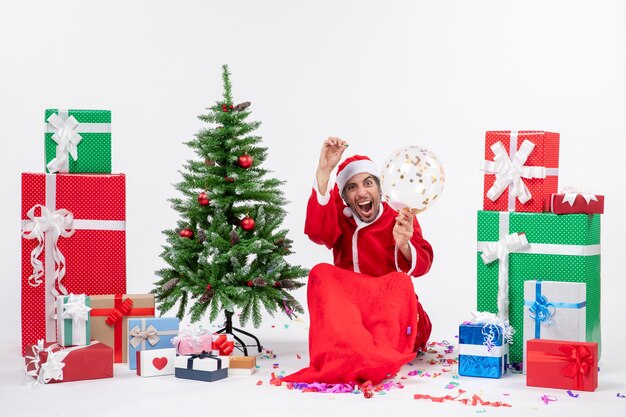 Humor navideño con jóvenes felices locas ha santa claus sentado cerca del árbol de navidad y regalos en diferentes colores sobre fondo blanco.