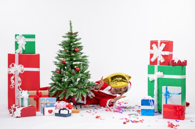 Foto gratuita humor navideño con santa claus acostado detrás del árbol de navidad cerca de regalos en diferentes colores sobre fondo blanco foto de stock