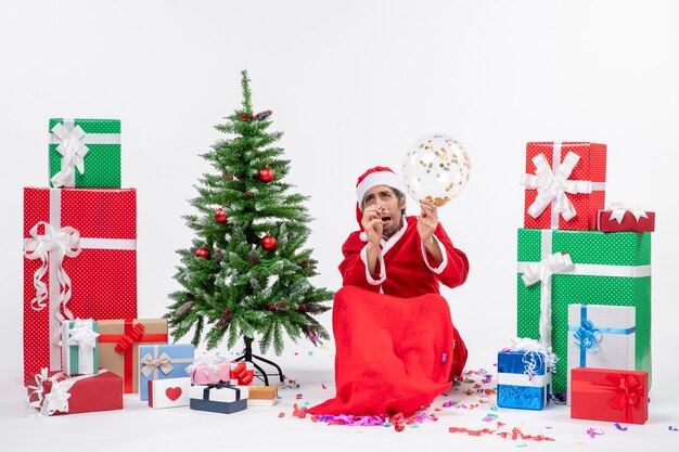 Humor navideño con santa claus asustado sosteniendo globo sentado cerca del árbol de navidad y regalos en diferentes colores sobre fondo blanco.