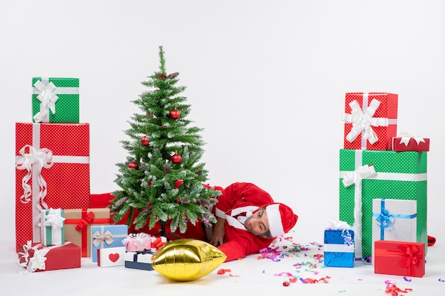 Humor navideño con santa claus cansado joven acostado detrás del árbol de navidad cerca de regalos en diferentes colores sobre fondo blanco.