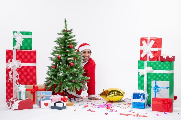 Humor navideño con santa claus joven escondido detrás del árbol de navidad cerca de regalos en diferentes colores sobre fondo blanco.