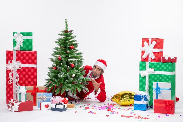 Humor navideño con santa claus joven escondido detrás del árbol de navidad cerca de regalos en diferentes colores sobre imágenes de fondo blanco