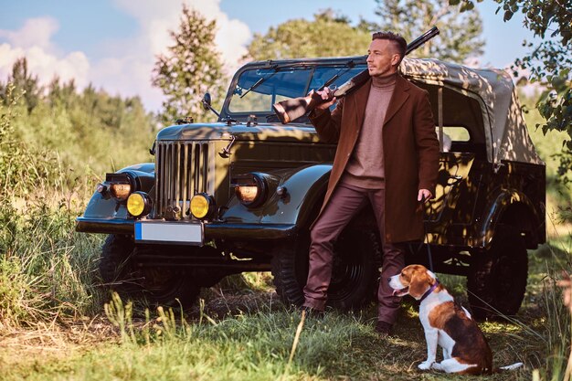 Hunter con ropa elegante sostiene una escopeta y está de pie junto a su perro beagle cerca de un auto militar retro en el bosque.