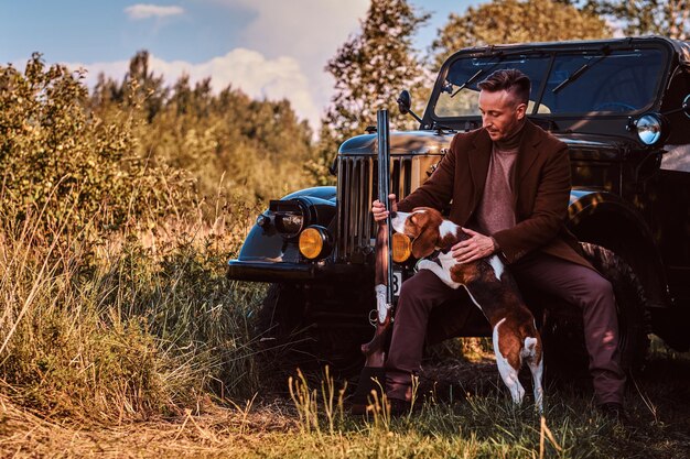 Hunter con ropa elegante sostiene una escopeta y se sienta junto con su perro beagle mientras se apoya en un auto militar retro en un bosque.