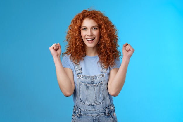 Hurra día exitoso. Los petos de verano de chica pelirroja cristiana alegre aprietan los puños con alegría sonriendo ampliamente determinado a lograr el éxito, triunfando el juego ganador de pie con un fondo azul optimista.