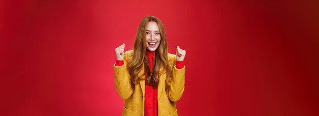 Foto gratuita hurra éxito retrato de feliz alegre y sincera linda jengibre mujer en abrigo amarillo levantando clenc