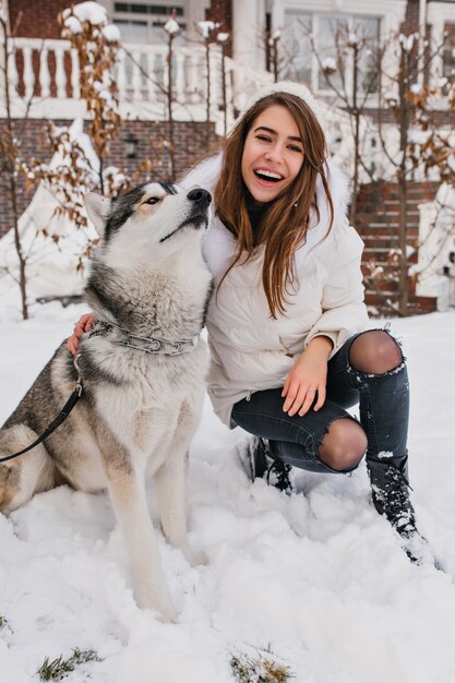 Husky divertido descansando después del juego en el parque cubierto de nieve. Retrato al aire libre de elegante mujer blanca en jeans rotos sentada en el suelo cerca de su hermoso perro en fin de semana de invierno.