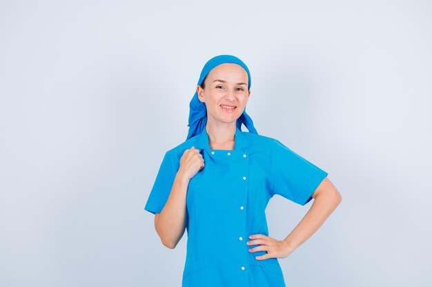 Identificación de enfermera sonriente mirando a la cámara sosteniendo la mano en el hombro y poniendo la otra mano en la cintura sobre fondo blanco