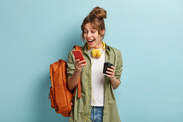 Imagen de una adolescente sonriente y alegre que disfruta de la comunicación con un amigo en el chat grupal, ve fotos divertidas en línea, escucha música sin conexión con auriculares, bebe café aromático de una taza de papel