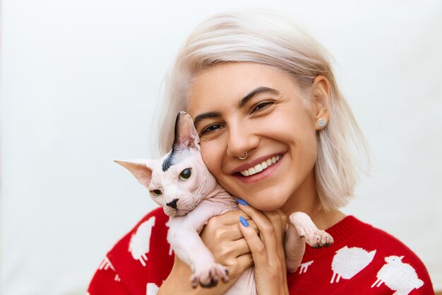 Imagen de adorable joven mujer caucásica vistiendo suéter de punto de moda abrazando a su linda mascota de raza alta. Gato Sphynx sin pelo ronroneando en brazos de su feliz dueña adolescente. Mascotas y personas