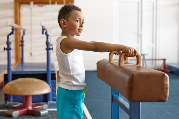 Imagen de adorable niño ateltico de piel oscura feliz en pantalones cortos azules y camiseta blanca posando en el gimnasio