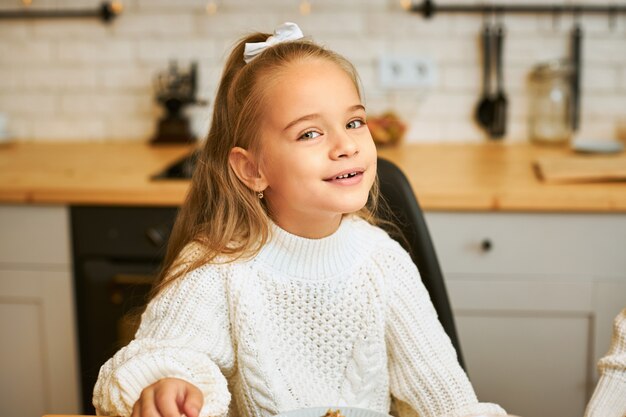 Imagen aislada de adorable niña con cinta blanca en el pelo posando en casa contra el interior de la cocina borrosa