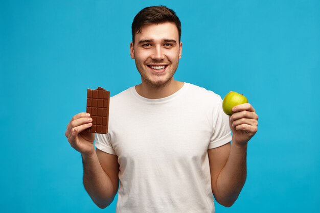 Imagen aislada de apuesto joven emocional manteniendo una estricta dieta libre de azúcar con manzana verde y barra de chocolate con leche, con expresión emocionada, yendo a comer alimentos prohibidos como comida trampa