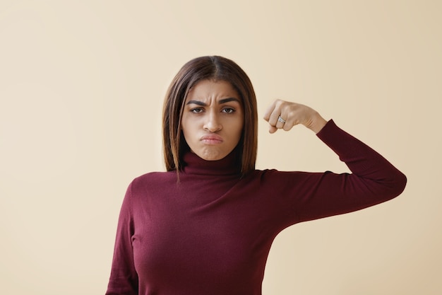 Imagen aislada de atractiva joven elegante mujer de piel oscura con cabello suelto con mirada furiosa loca, haciendo muecas y sosteniendo el puño bombeado frente a ella, lista para golpear. Feminismo y poder femenino