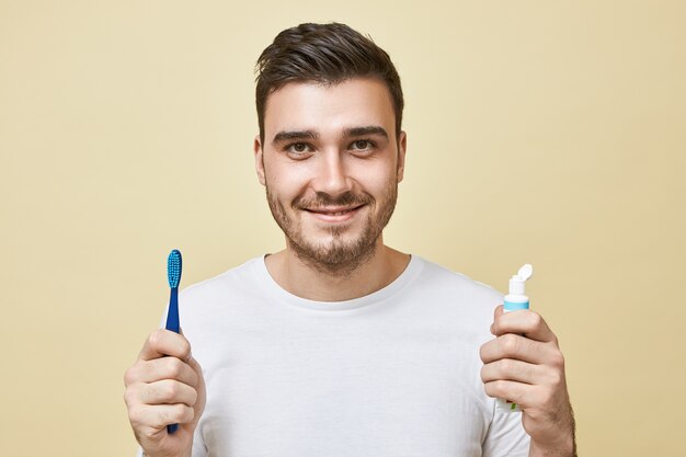 Imagen aislada de confiado alegre joven morena con cerdas sosteniendo cepillo y tubo de pasta de dientes, cepillándose los dientes justo después de despertar. Concepto de higiene, rutina matutina y blanqueamiento dental.