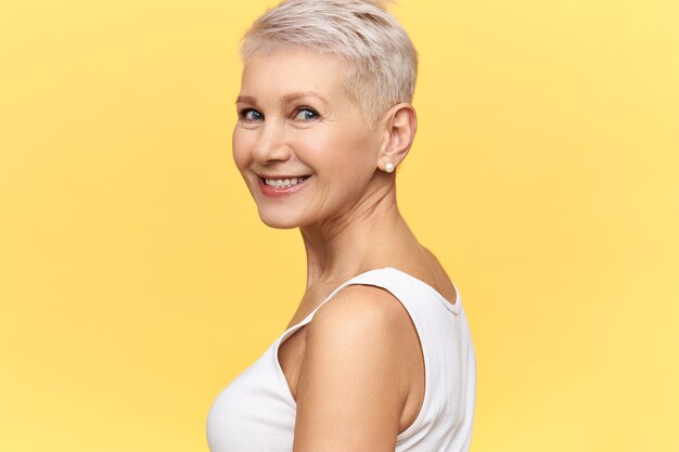 Imagen aislada de la hermosa y encantadora mujer de mediana edad con cabello corto teñido girando la cabeza, sonriendo felizmente, posando sobre fondo amarillo en blanco con espacio de copia