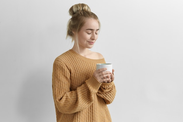 Imagen aislada de hermosa mujer joven encantadora con un nudo de pelo sosteniendo una taza grande disfrutando de un capuchino recién hecho de una taza grande, vistiendo un acogedor jersey de gran tamaño, manteniendo los ojos cerrados y sonriendo