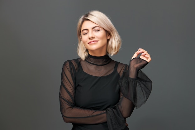 Imagen aislada de hermosa mujer joven con estilo vistiendo elegante vestido de noche y anillo en la nariz posando con los ojos cerrados, sonriendo, disfrutando de la agradable música festiva. concepto de invierno y vacaciones