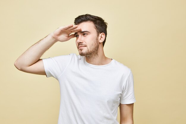 Imagen aislada de hombre joven guapo con barba de dos días sosteniendo la mano en la frente y entrecerrando los ojos, mirando pensativamente a la distancia, tratando de ver algo lejano, teniendo problemas de visión