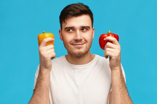 Imagen aislada de un joven atractivo lindo que tiene una expresión facial misteriosa pensativa, mirando a otro lado, sosteniendo dos pimientos, pensando qué cocinar para una cena vegetariana o comparando