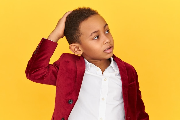 Imagen aislada de lindo colegial afroamericano confundido mirando hacia arriba con expresión facial perpleja perpleja rascándose la cabeza, se olvidó de hacer la tarea, avergonzado.