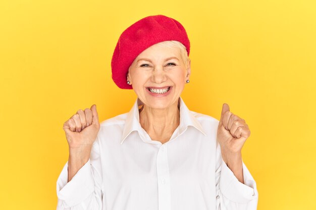 Imagen aislada de la mujer francesa madura alegre positiva de moda con camisa blanca y gorro rojo manteniendo los puños apretados y sonriendo ampliamente, llena de alegría con buenas noticias, gritando Sí con entusiasmo