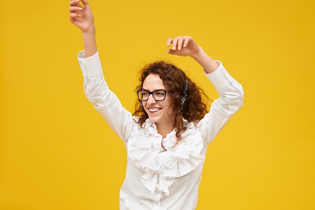 Imagen aislada de mujer joven emocional positiva con cabello oscuro y rizado posando en la pared amarilla con las manos en el aire, bailando, escuchando música en auriculares, sonriendo con entusiasmo, con gafas