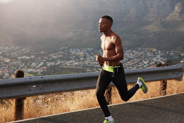 La imagen al aire libre del deportista corre en una carretera de montaña rural, fotografiada en movimiento, tiene una forma de cuerpo atlético, entrena trotando durante el clima cálido, entrena resistencia, intenta no detenerse para descansar