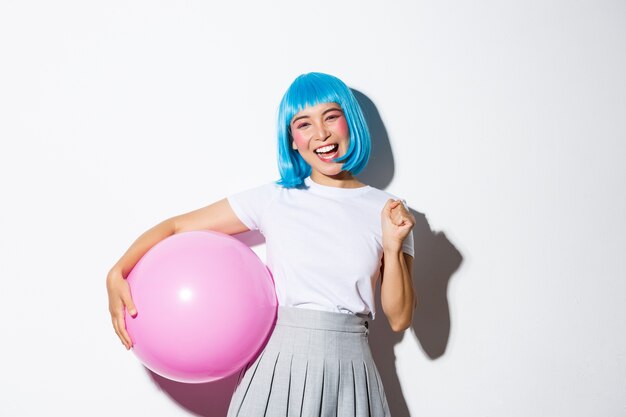Imagen de una alegre chica asiática ganadora, luciendo feliz y triunfante, celebrando las vacaciones, vistiendo traje de fiesta y peluca azul