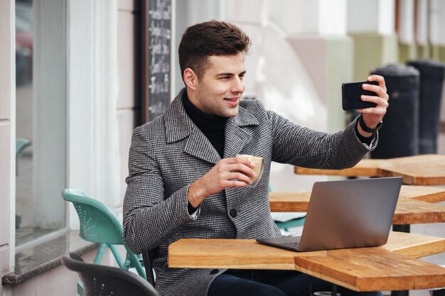 Imagen de alegre hombre morena haciendo selfie o skyping mientras descansa en la cafetería de la calle y tomando café de vidrio