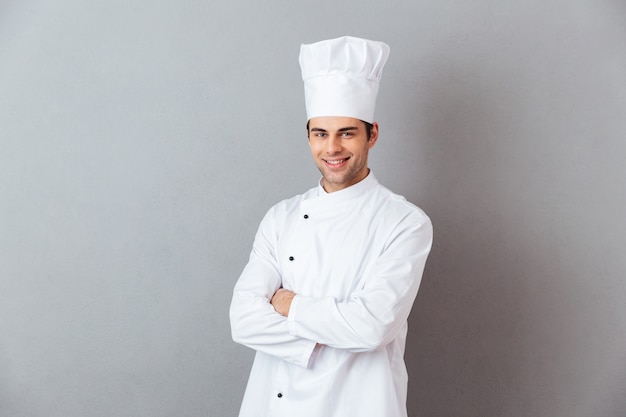 Imagen de alegre joven cocinero en uniforme de pie aislado sobre pared gris