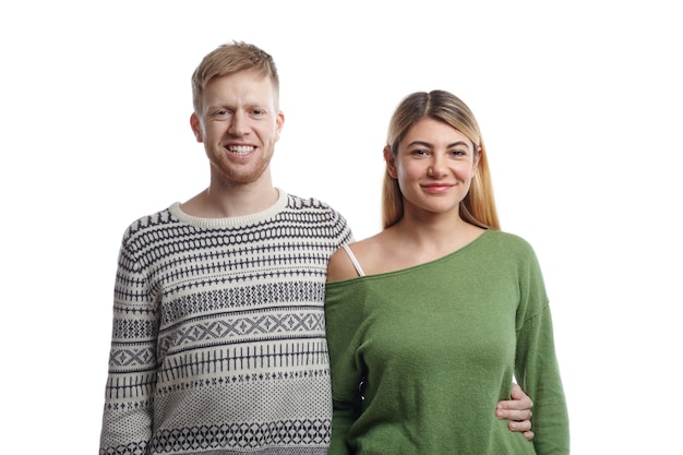 Imagen de la alegre pareja europea joven en ropa elegante posando con sonrisas felices: chico barbudo en suéter abrazando a su novia rubia por la cintura. Personas, amor y relaciones